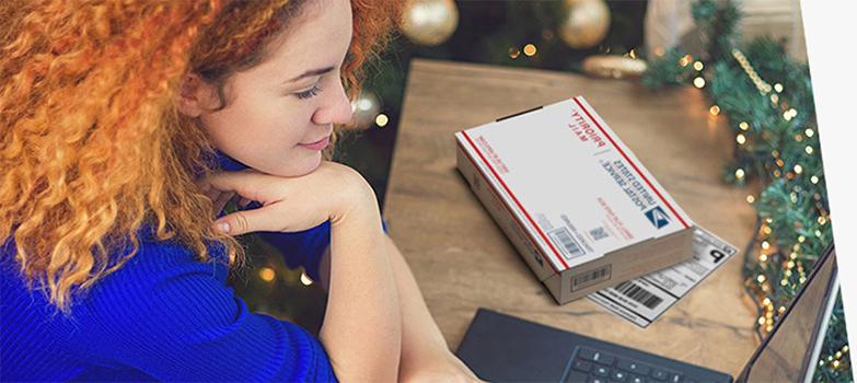 Woman on laptop with Priority Mail box and printed shipping label on the table next to her.